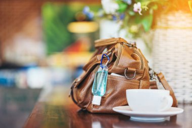 Corona Virüsü 'nü (Covid-19) öldürmek için mini alkol jeli şişesi kafede üzerinde sıcak kahve olan kahverengi deri bir omuz çantasına asılır. Yeni normal yaşam tarzı. Sağlık hizmetleri kavramı. Alkol jeline odaklanın.