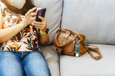 Corona Virüsü 'nü (Covid-19) öldürmek için mini taşınabilir alkol jeli şişesi kahve dükkanındaki koltukta kahverengi deri bir omuz çantasına asılmış. Yeni normal yaşam tarzı. Sağlık hizmeti konsepti. Alkol jeline odaklan