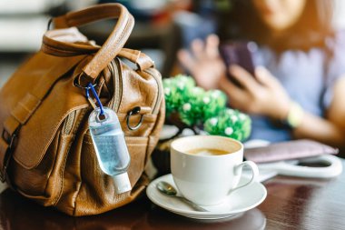 Corona Virüsü 'nü (Covid-19) öldürmek için mini alkol jeli şişesi kafede üzerinde sıcak kahve olan kahverengi deri bir omuz çantasına asılır. Yeni normal yaşam tarzı. Sağlık hizmetleri kavramı. Alkol jeline odaklanın.