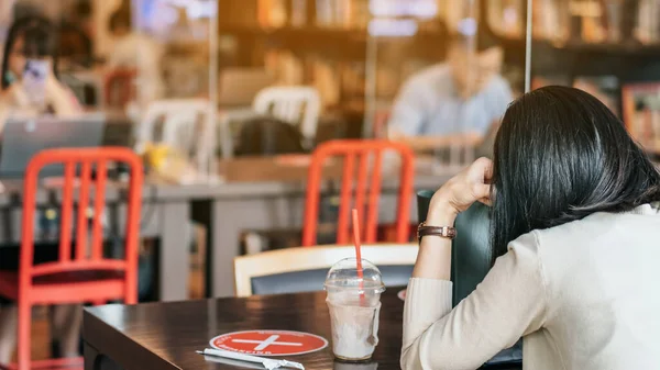 Back view of Asian woman use smartphone and relax with beverage in coffee shop, where seats are spaced for social distancing during the Corona virus (COVID-19) epidemic. New normal lifestyle