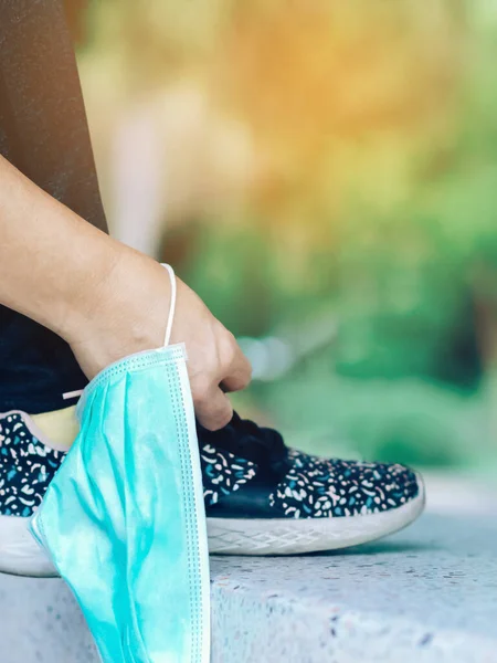 Fitness woman take off her surgical mask getting ready to do run exercise lacing running shoes for outdoor jogging. New normal lifestyle. Health care concept. Selective focus on surgical mask.