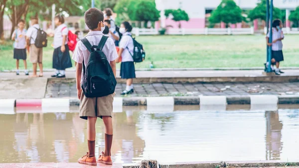 Male elementary school student wear face mask to prevent the Coronavirus(Covid-19) wait for her parents to pick her up to return home after school and the rain just stop in front of the school gate