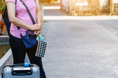 Elinde cüzdanı ve sırt çantasıyla yol kenarında otobüs bekleyen kadın bir gezgin. Koronavirüs konsepti sırasında güvenlik yolculuğu. Yeni normal yolcu. Cerrahi maskeye odaklan.