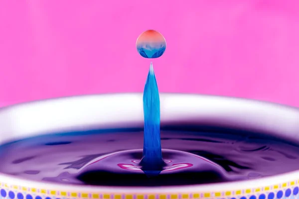 A blue and orange water drop on a pink background. Orange coloured milk dropped into blue liquid.