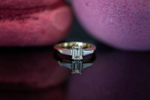 An emerald cut diamond and two side diamonds set into a yellow gold ring and reflected on a shiny black surface
