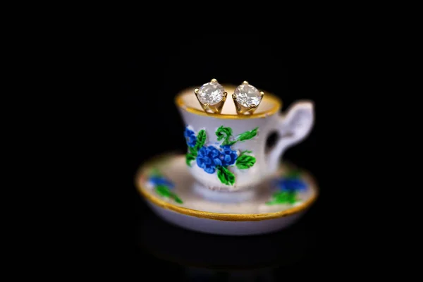 A pair of diamond stud earrings set in yellow gold, sitting in a miniature floral teacup against a black background