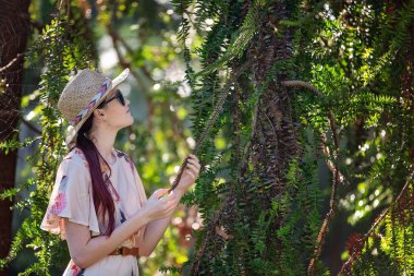 Hasır şapka, güneş gözlüğü ve dikenli bir yaprak inceleyerek çiçek elbise giyiyor uzun kızıl saçlı genç ve güzel kadın