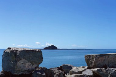 Liman duvarındaki dev kayalar ufuktaki küçük bir ada, parlak mavi bir okyanusla çevrili.
