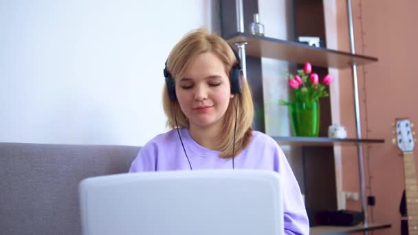 En flicka sitter på en soffa med hörlurar lyssnar på musik med hörlurar och fungerar i en bärbar dator. — Stockvideo