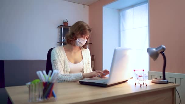 A lány otthon dolgozik a karantén alatt. Coronavirus-járvány. Egy lány sebészeti maszkban egy laptopon dolgozik. Távoli munka. — Stock videók