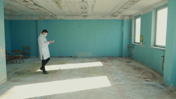 Un trabajador médico con traje protector y gafas camina a través de una habitación desierta mirando las líneas y escribiendo datos en una carpeta . — Vídeos de Stock