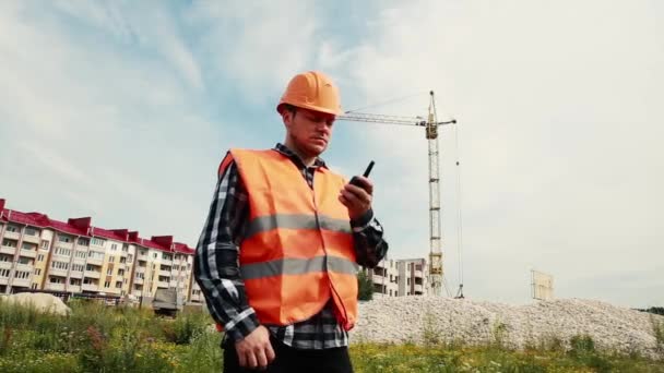 Um construtor masculino falando em um walkie-talkie no fundo de um canteiro de obras . — Vídeo de Stock