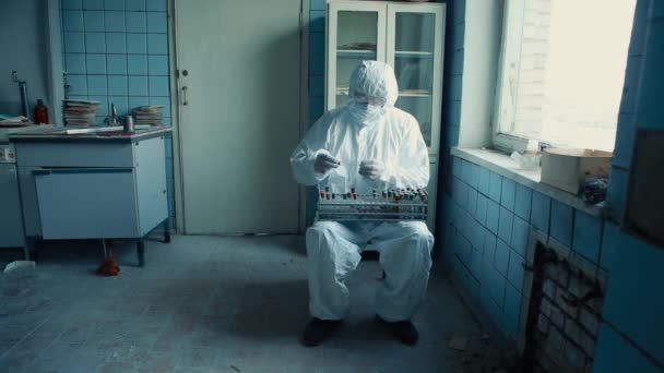 Ο ιατρός με προστατευτική στολή και τα γυαλιά κάθεται σε μια καρέκλα στο εργαστήριο εξετάζει τις φιάλες με αναλύσεις. — Αρχείο Βίντεο