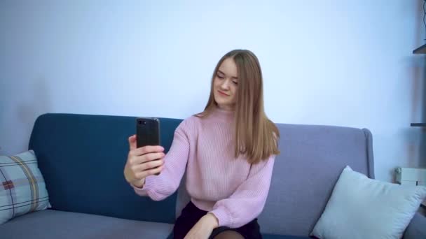 Een meisje in een grote kamer zit op een bank en fotografeert zichzelf aan de telefoon. — Stockvideo