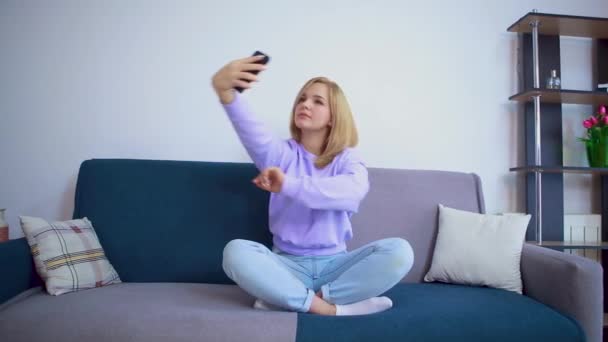 Vacker blond flicka tar en selfie medan du sitter på en soffa. — Stockvideo