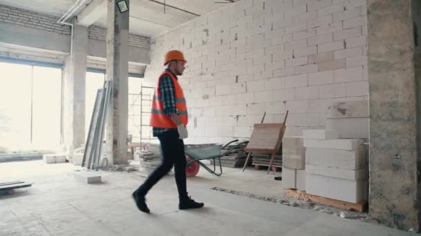 Мужчина-строитель в оранжевом жилете и оранжевом шлеме подходит к стопке кирпичей, выбирает и поднимает один и оставляет . — стоковое видео