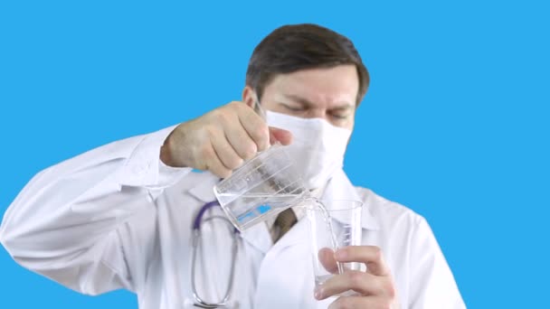 En man i medicinsk rock och mask häller vatten från ett kärl i ett annat. — Stockvideo