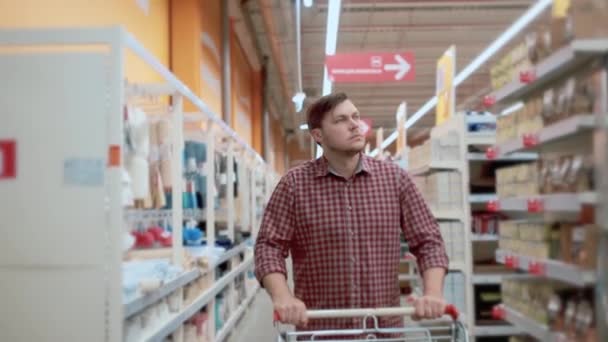 Een man met een trolley in een winkel gaat langs de planken met goederen. — Stockvideo