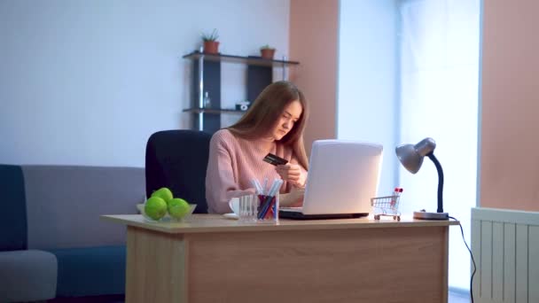 Een meisje werkt op een laptop zittend aan een tafel op afstand, voert gegevens van een creditcard in. — Stockvideo