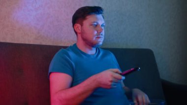 Kanepede elinde uzaktan kumandayla oturan bir adam cips yiyor ve TV 'de duygusal kanalları değiştiriyor..