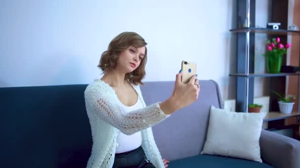 Piękna dziewczyna siedzi na kanapie robi selfie. — Wideo stockowe