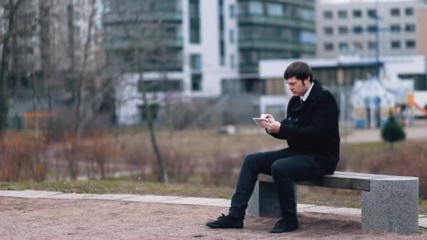 Un hombre de negocios con un abrigo negro está al aire libre sentado en un banco y trabajando en una tableta . — Vídeos de Stock