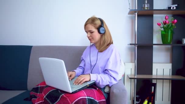 A girl on a sofa and in headphones wrapped in a checkered plaid communicates via video link on a laptop. — Stock Video