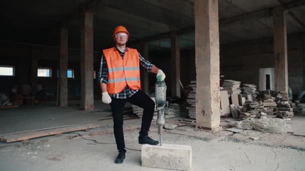 Ritratto di un muratore con giubbotto arancione e casco arancione sullo sfondo di un cantiere che tiene in mano un martello pneumatico . — Video Stock