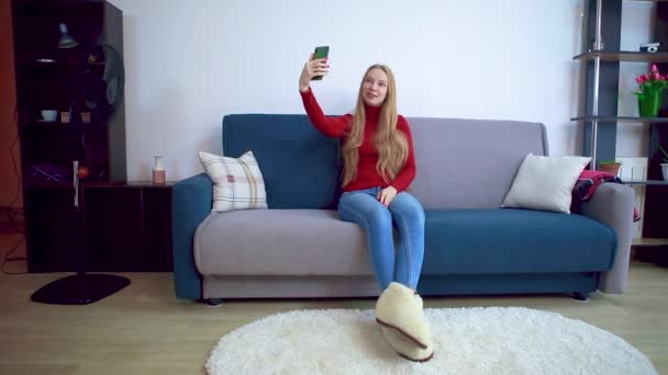 Mladá dívka v červeném svetru sedí na pohovce se selfie. — Stock video