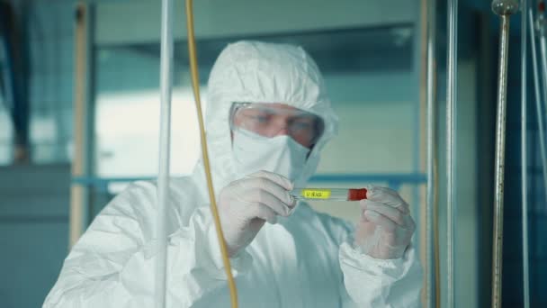 Un asistente de laboratorio masculino con traje protector y máscara sostiene un tubo de ensayo con el virus COVID-19 en sus manos . — Vídeos de Stock