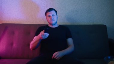 Kanepede oturan bir adam elinde uzaktan kumandayla televizyon kanallarını yavaşça değiştiriyor..
