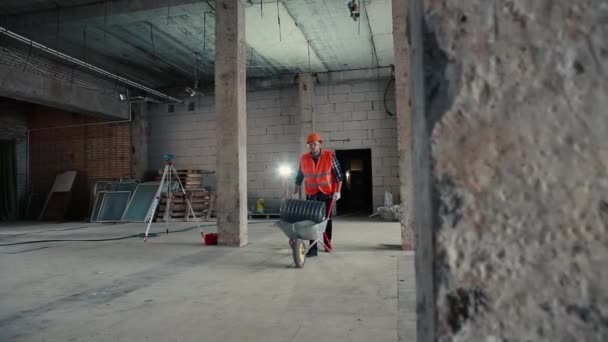 Un constructor masculino con chaleco naranja y casco naranja lleva un carro de construcción sobre un fondo de obra . — Vídeos de Stock