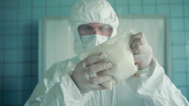 De laboratoriumassistent in een beschermend pak en bril houdt in zijn handen een zak met wit poeder in zijn handen.. — Stockvideo