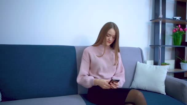 En ung flicka sitter på en soffa surfar i en smartphone. — Stockvideo