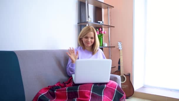 一位年轻的金发女子坐在沙发上，通过笔记本电脑上的视频交流进行交流. — 图库视频影像