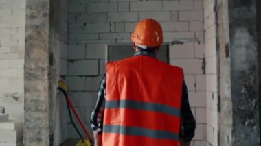 Turuncu yelek ve turuncu kasklı bir erkek müteahhit yapım aşamasındaki bir nesneye geri dönüyor..