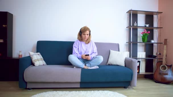 Дівчина на дивані і в навушниках сидить на дивані і слухає музику . — стокове відео