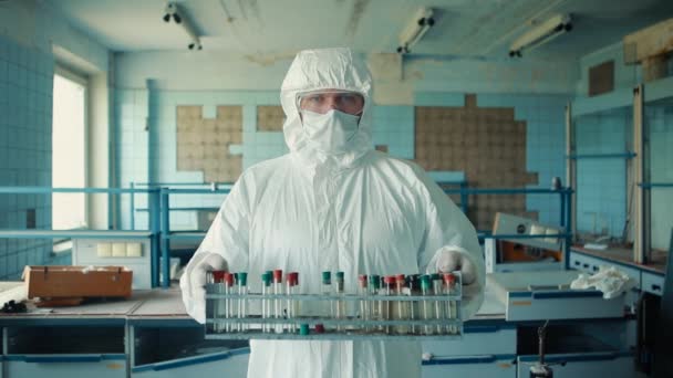 Porträt eines medizinischen Mitarbeiters in Anzug und medizinischer Maske hält in seinen Händen eine Schachtel mit Reagenzgläsern auf dem Hintergrund eines zerstörten Labors. — Stockvideo