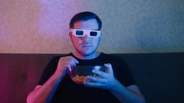 Egy férfi sztereó szemüvegben ül egy kanapén, chipset eszik egy foltos tányérról.. — Stock videók