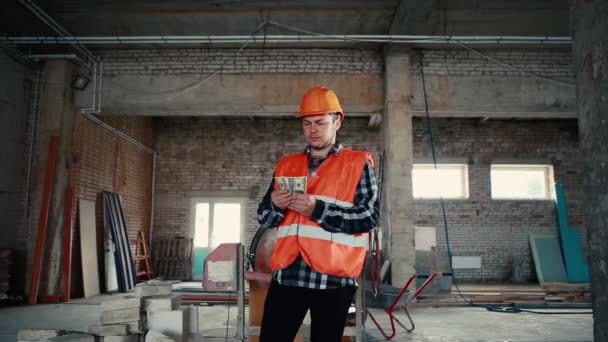 A male builder is standing in a building amid a construction site and is slowly recounting money. — Stock Video