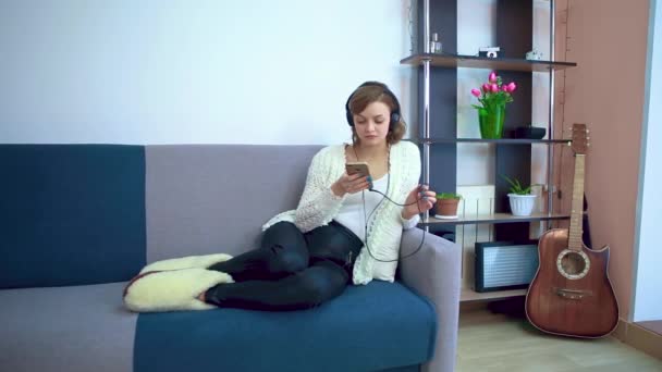 A young girl in the living room listens to music on headphones from a smartphone. Cozy home interior. The concept of a happy lifestyle. — Stock Video