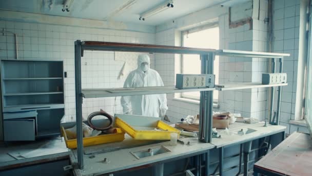 En man i skyddsdräkt och skyddsglasögon passar i ett förstört laboratorium. — Stockvideo