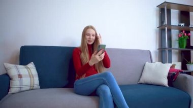 Kırmızı kazaklı bir kız kanepede oturmuş akıllı telefondan konuşuyor..