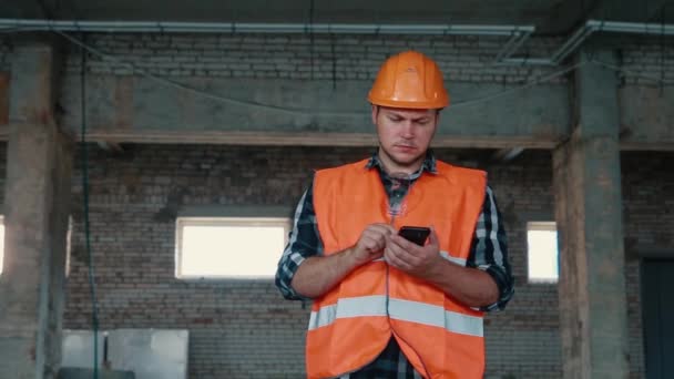 オレンジ色のベストとオレンジのヘルメットの男性ビルダーは、スマートフォン上でデータを書き込み、文書化します. — ストック動画