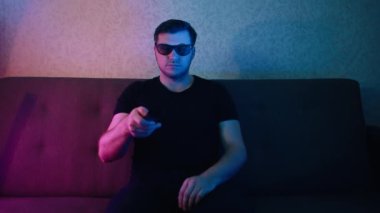 Bir adam kanepede oturup 3 boyutlu televizyon izliyor ve uzaktan kumandayla TV 'yi değiştiriyor..