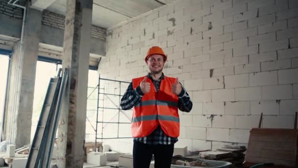 Un constructor masculino con un chaleco naranja y un casco naranja está de pie en una habitación en medio de un sitio de construcción muestra alegremente con sus manos que todo está bien . — Vídeos de Stock