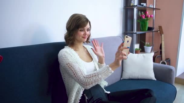 Girl talking on a video call on a smartphone, sitting on a gray sofa. — Stock Video