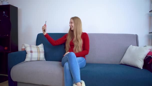 Een meisje in een rode trui zittend op een bank neemt een selfie. — Stockvideo