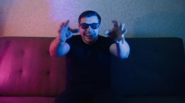 Üç boyutlu gözlüklü bir adam kanepede oturur ve TV 'de ilginç bir sahne izler, elleriyle farklı hareketler yapar..