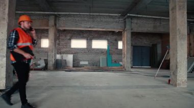 Turuncu yelek ve turuncu kasklı bir erkek müteahhit inşaat alanında yürüyor, telefonla konuşuyor..